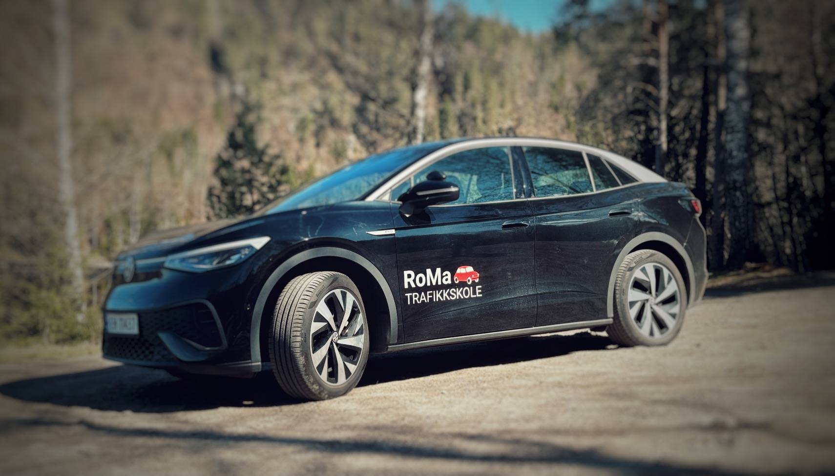 Svart elbil fra RoMa Trafikkskole med logoen på døra, parkert på en grusplass foran skogen